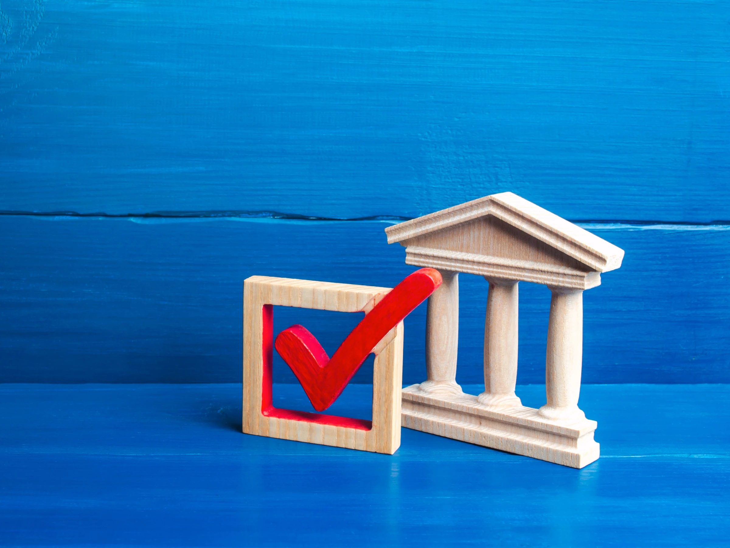 Wooden check mark symbol next to a wooden courthouse model, representing government approval or compliance, on a blue background.