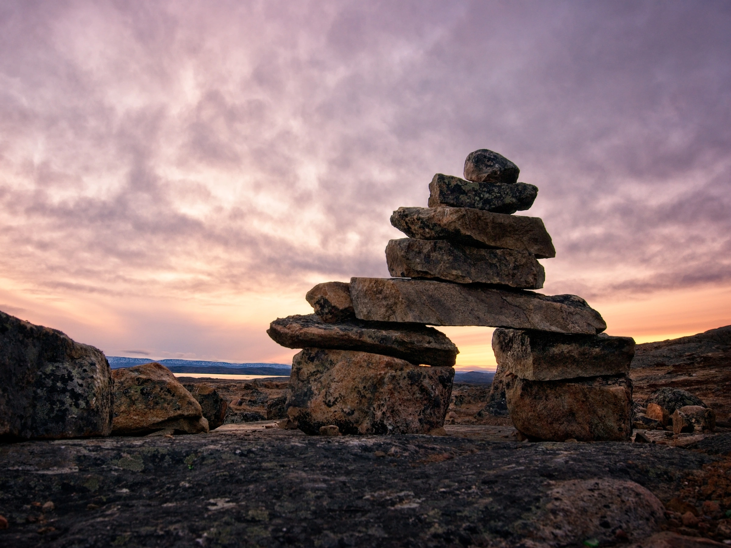Stone inuksuk silhouette against a colorful sunset sky in a rocky Arctic landscape.