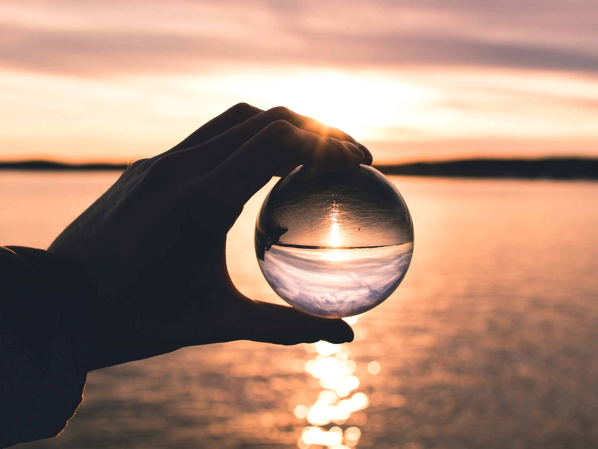 Main tenant une sphère en verre reflétant à l’envers un coucher de soleil sur une étendue d’eau calme.